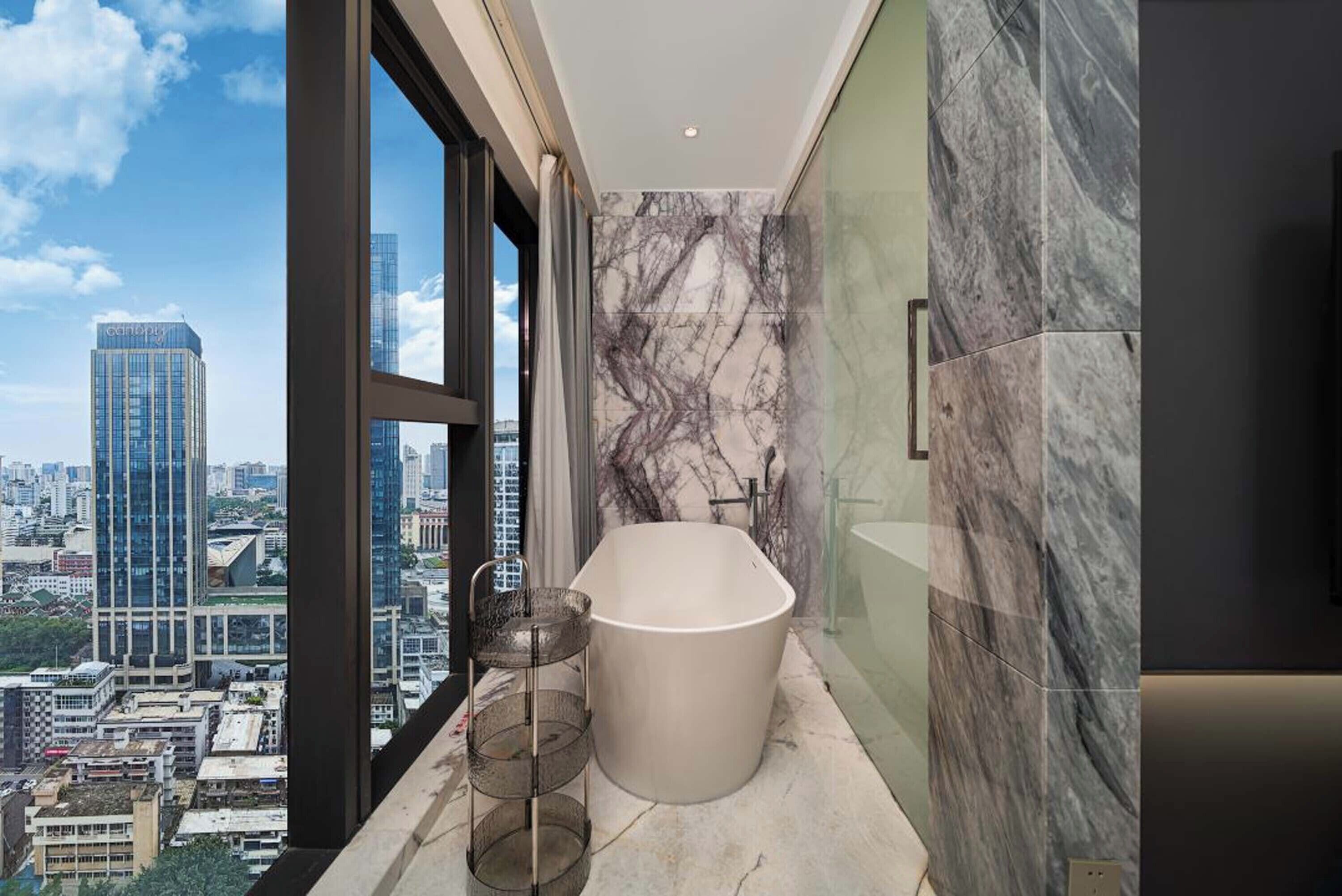 Un baño moderno con una bañera independiente, paredes de mármol y vistas a la ciudad a través de amplios ventanales.