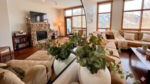 Una sala de estar con chimenea de piedra, televisor, sofá y ventanas amplias con vistas a las montañas.