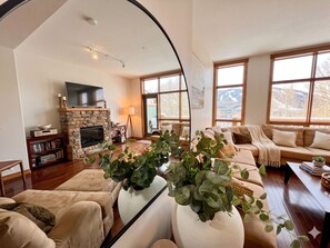 A living room with a stone fireplace, a television, a sofa, and large windows with a view of mountains. - Your 4-Season Adventure Base! True ski-in/ski-out 4BR condo at Sullivan Stone. Prime location on the Main Run. Features a private hot tub and a gourmet kitchen for the ultimate mountain getaway. (Kimberley)