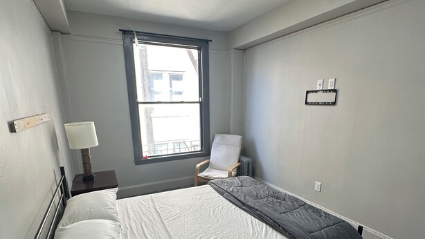 Ein Schlafzimmer mit einem Bett, einem Sessel, einem Nachttisch, einer Lampe, einem Fenster und einer Wandleuchte.