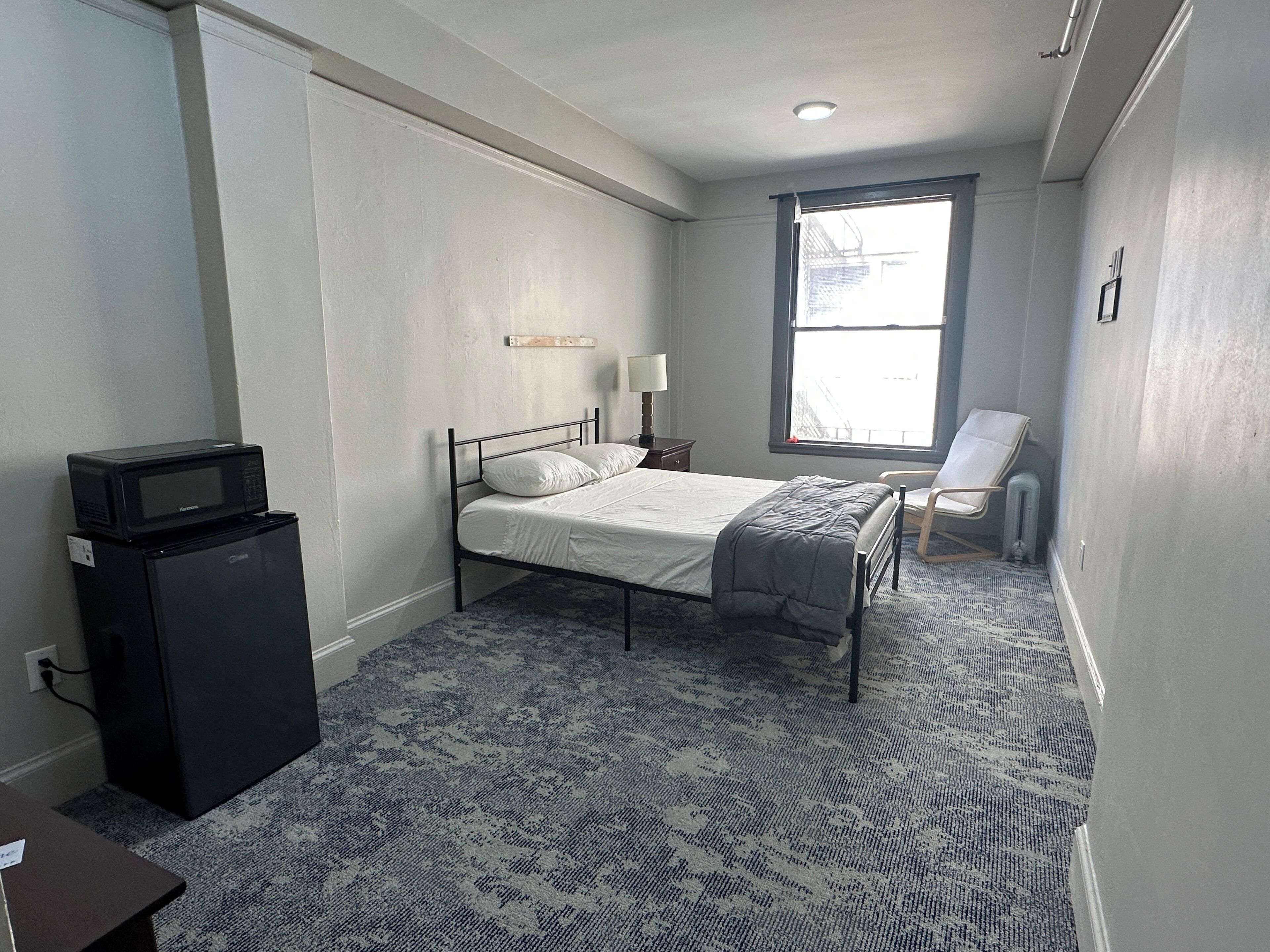 A bedroom with a bed, a desk, a chair, a window, and a microwave.