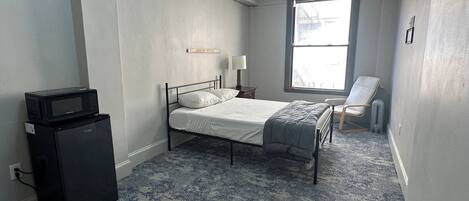 A bedroom with a bed, a desk, a chair, a window, and a microwave.