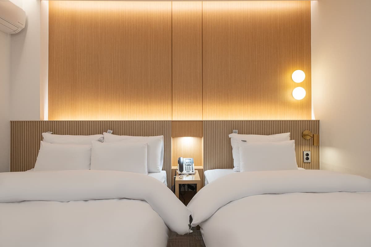 Habitación de hotel con dos camas, un cabecero de madera, una mesita de noche con teléfono y una lámpara de pared.