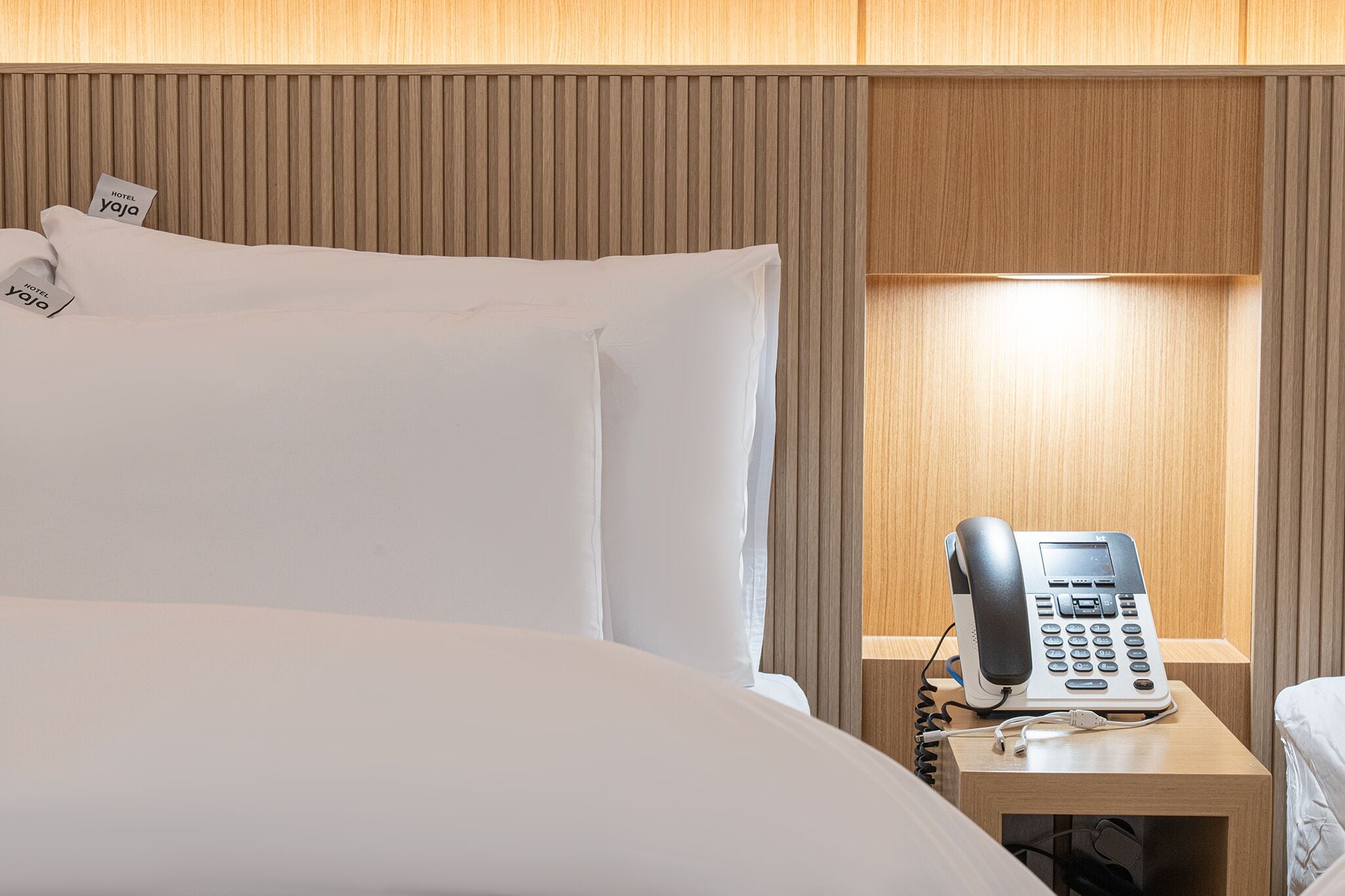 A hotel room with a bed, pillows, a bedside table with a telephone, and a wooden headboard.