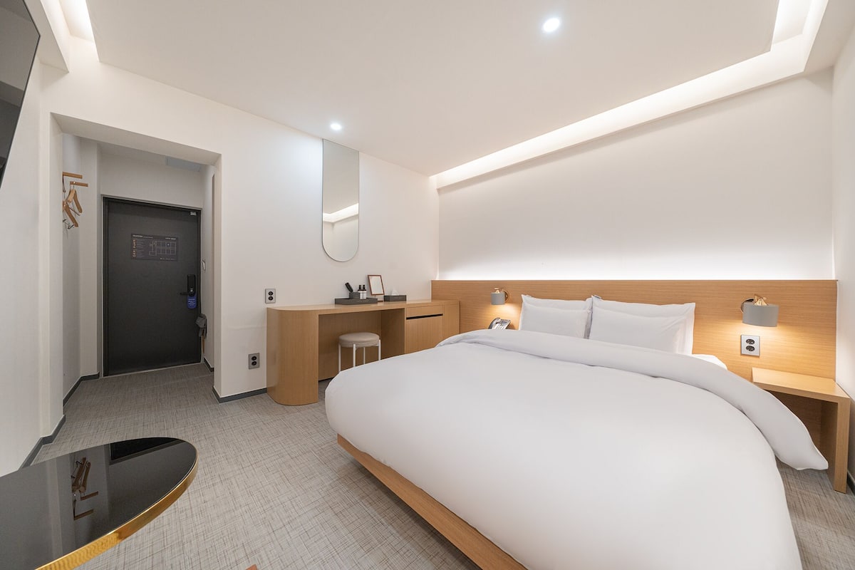 Una habitación de hotel moderna con una cama grande, un escritorio, una silla, un espejo y una mesita de noche con lámpara.