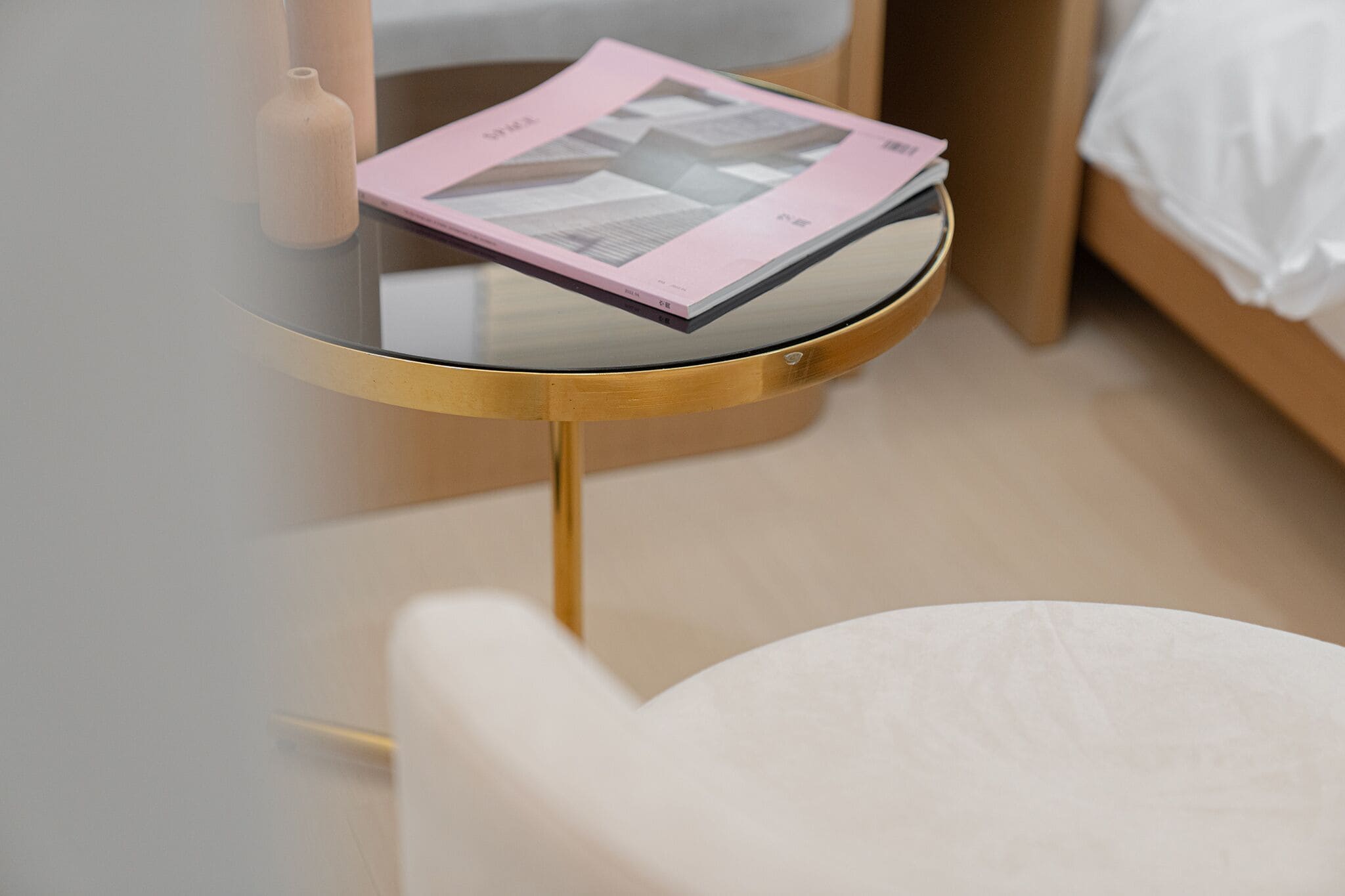 A round glass table with a gold base, a pink book, and a small wooden vase.