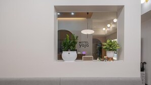 Un área de recepción de hotel moderna con una paleta de colores neutros, que incluye un mostrador de recepción, plantas en macetas y un letrero que dice "HOTEL YAJI".
