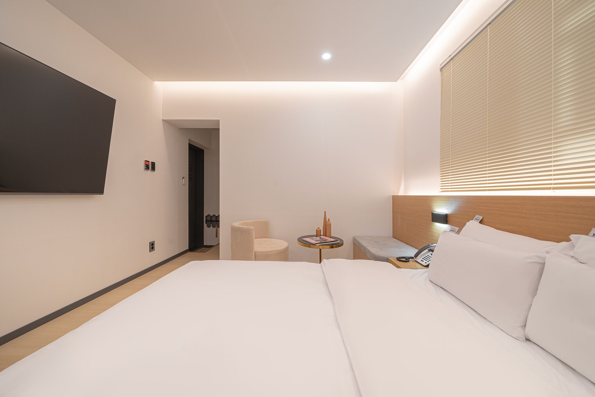A modern hotel room with a large bed, a bedside table with a lamp, a chair, and a TV mounted on the wall.