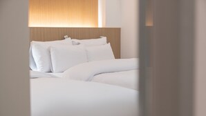 Una cama con ropa de cama y almohadas blancas, cabecera de madera y lámpara de pared.