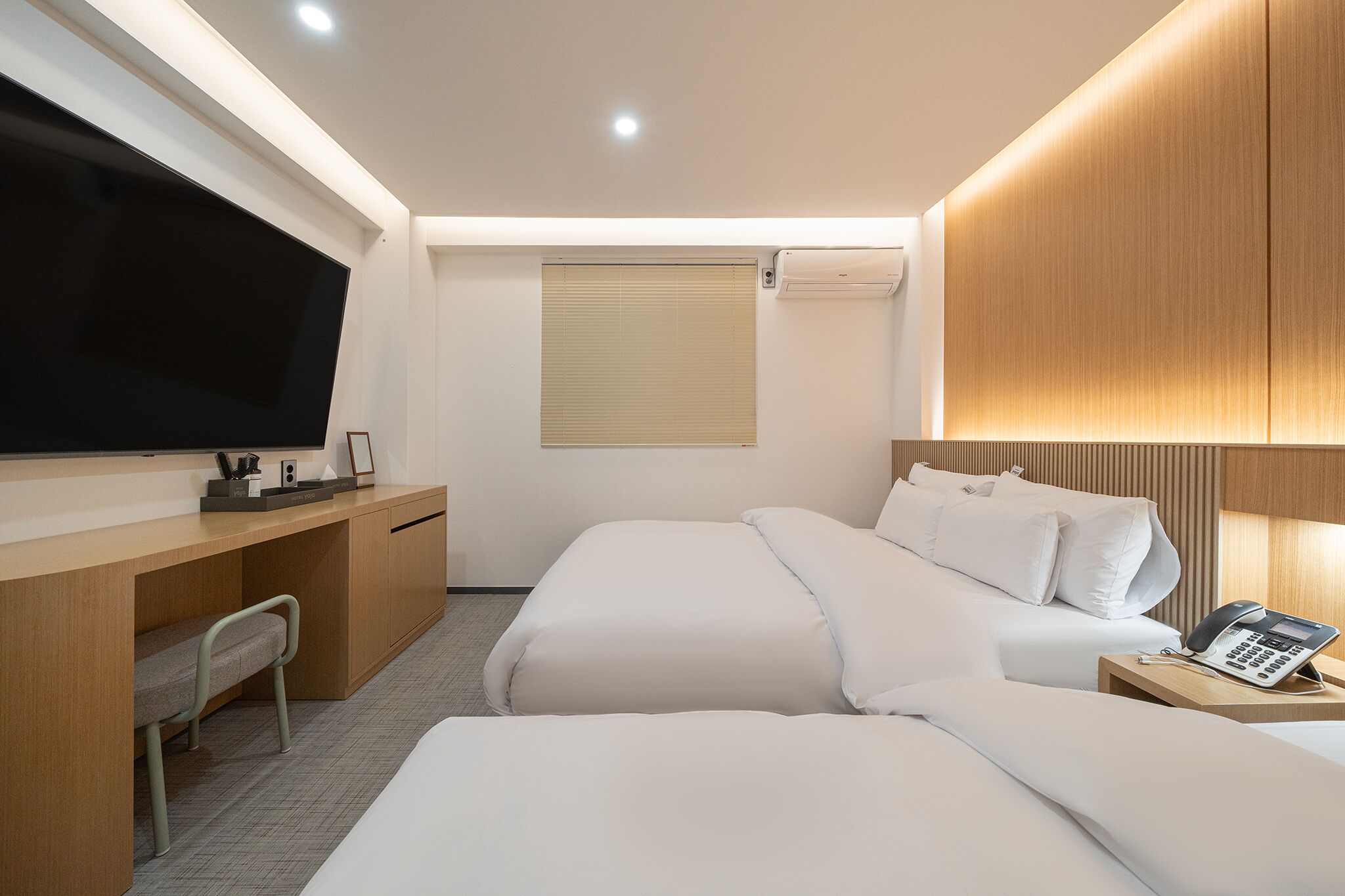 A hotel room with a large flat-screen TV, a desk with a chair, two beds, a bedside phone, and a window with blinds.