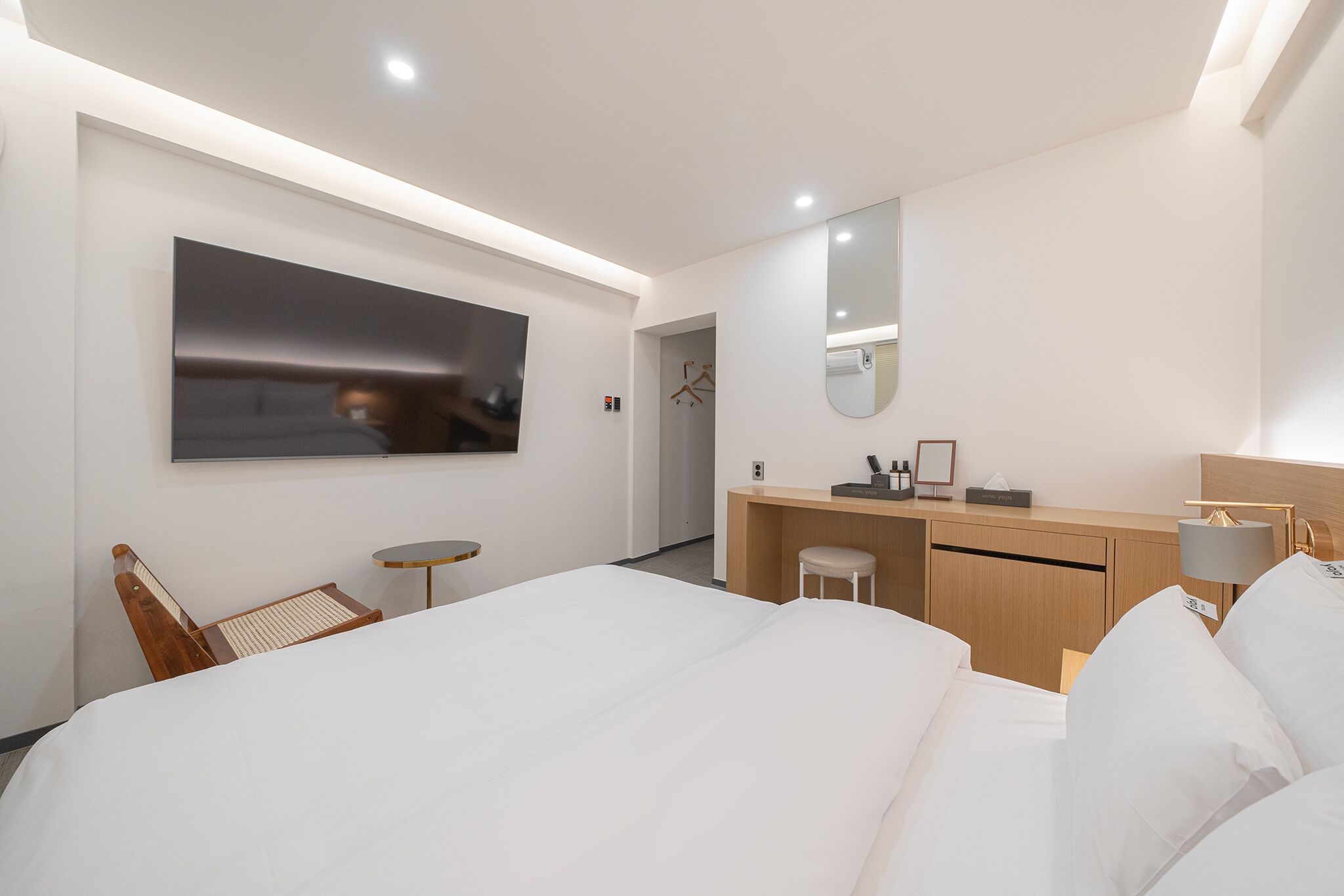 A modern hotel room with a large bed, a desk, a chair, a small table, a mirror, and a wall-mounted TV.