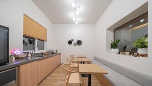 Una cocina moderna con armarios de madera, horno empotrado y encimera con cafetera. Hay dos mesas y sillas de madera, y una ventana con persianas.
