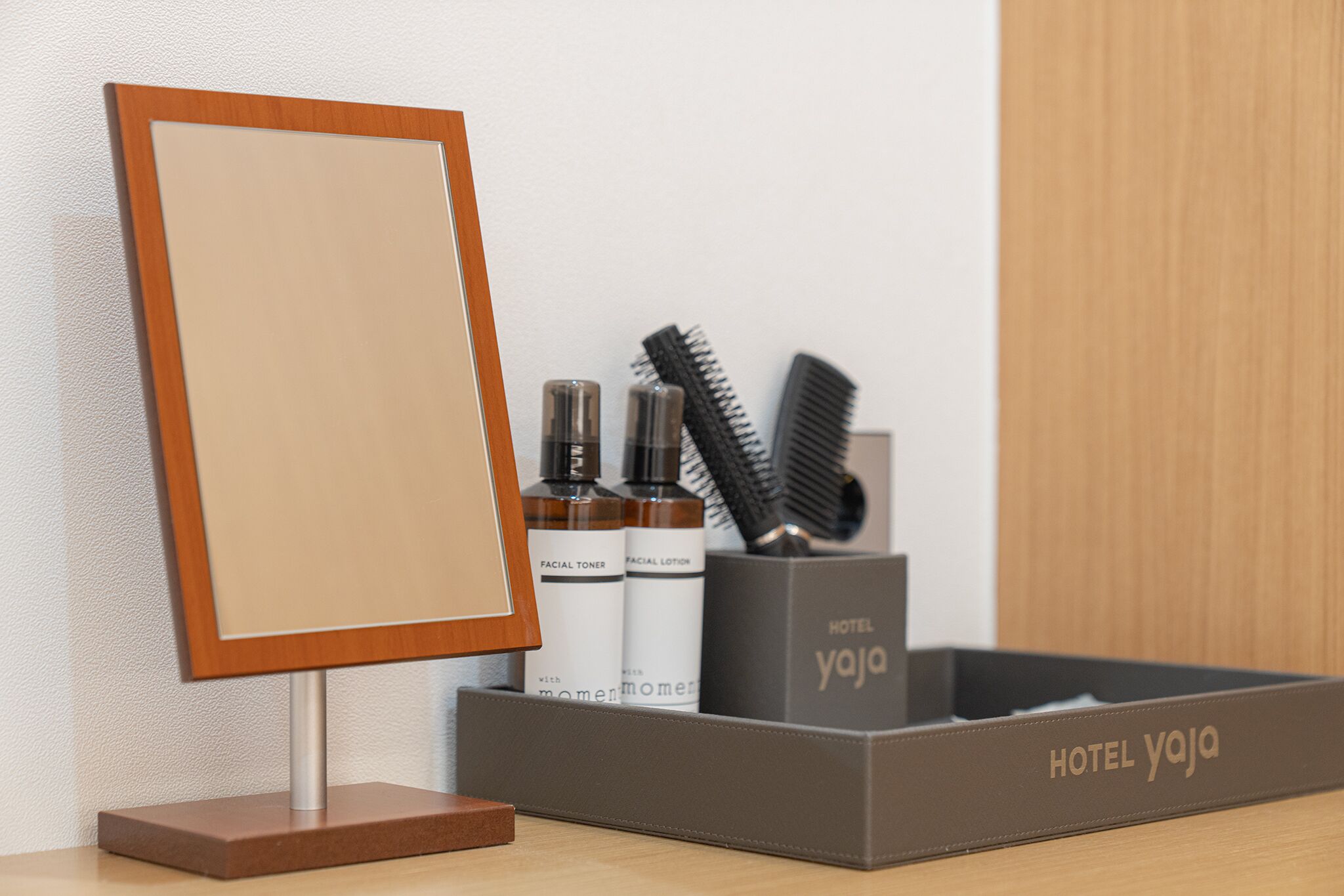 A hotel room with a wooden-framed mirror, a tray with toiletries including facial toner and lotion, and a black comb.