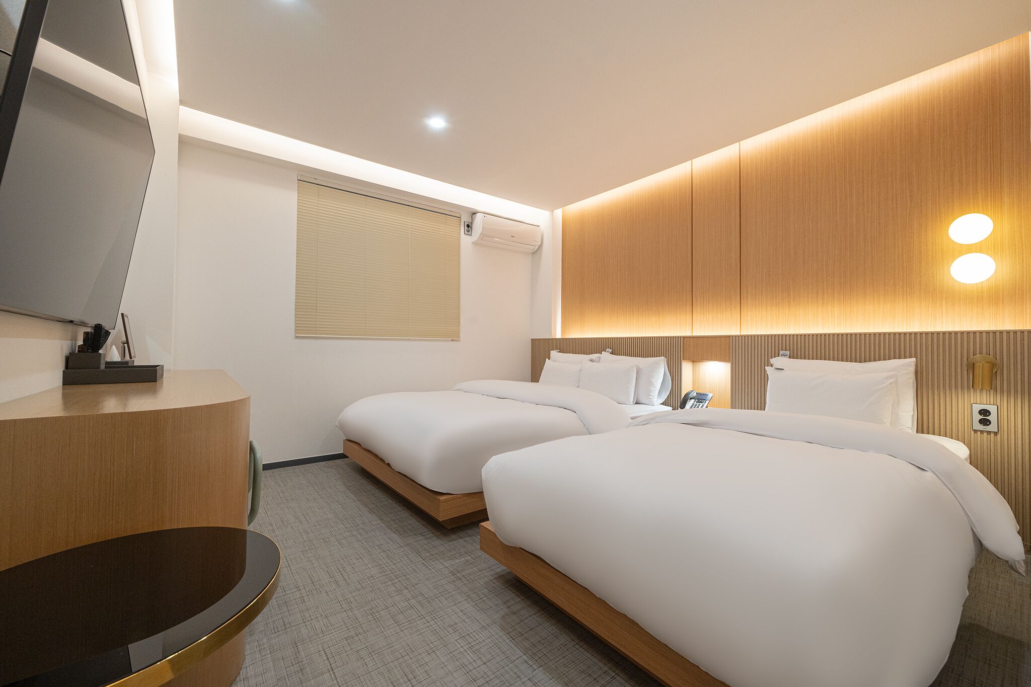 A hotel room with two beds, a desk, a TV, and a wall-mounted light fixture.