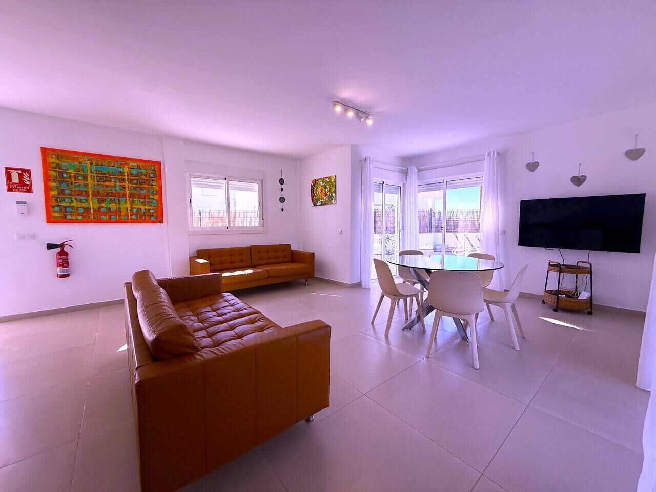 Una sala de estar moderna con un sofá marrón, una mesa de comedor con sillas, un televisor de pantalla plana y ventanas amplias.