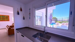 A modern kitchen with a large window, a built-in sink, and a view of a pool and lounge chairs. - Villa Modern Princess (Golf Del Sur)