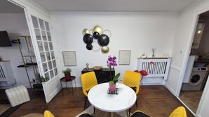 A compact living space with a dining area featuring a round table and yellow chairs with black cushions. A potted plant and a small table with a lamp are on the dining table. The room has a white door, a radiator, and a washing machine in the background. - House with fenced exterior and garage 2 steps from the spa. 2 adults/1 child  (Salies-de-Béarn)