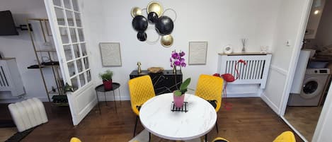 A compact living space with a dining area featuring a round table and yellow chairs with black cushions. A potted plant and a small table with a lamp are on the dining table. The room has a white door, a radiator, and a washing machine in the background.