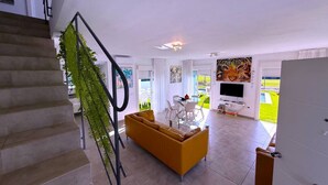 A modern living room with a sofa, coffee table, and TV. A staircase with a railing is visible on the left. - Villa 6- Pink Princess (Golf Del Sur)