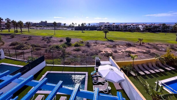 A pool area with lounge chairs and umbrellas, a grassy field, and a clear view of the ocean. - Villa 6- Pink Princess (Golf Del Sur)
