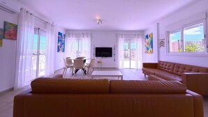 Una sala de estar moderna con un sofá grande, una mesa de comedor con sillas, un televisor de pantalla plana y ventanas amplias con cortinas.