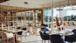 A modern dining area with tables set for a meal, large windows offering a view of the city, and a decorative plant centerpiece. - Status Apartments 100 (Becici)