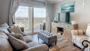 Una sala de estar con sofá, reposapiés y mueble para televisión, un balcón con cortinas y un ventilador de techo.