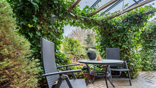 Eine Terrasse im Freien mit einem runden Tisch und vier Stühlen, umgeben von üppigem Grün und einer Pergola.