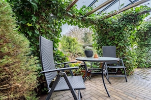 Um pátio externo com uma mesa redonda e quatro cadeiras, cercado por vegetação exuberante e uma estrutura de pérgola.