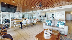 Amplia sala de estar con techo de madera, televisor de pantalla plana, comedor con sillas blancas y sofá con cojines estampados.