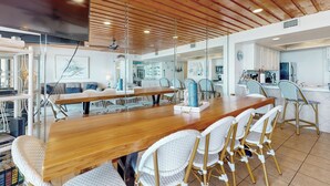 Un comedor amplio con una larga mesa de madera y sillas de mimbre blanco, una zona de cocina con armarios blancos y un techo moderno con vigas de madera.