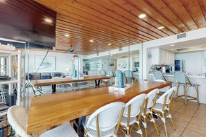 Ein geräumiger Essbereich mit einem langen Holztisch und weißen Korbstühlen, ein Küchenbereich mit weißen Schränken und eine moderne Decke mit Holzbalken.