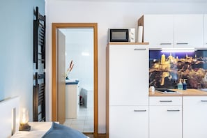 Habitación compacta con cama, escritorio con lámpara, microondas y área de cocina con armarios y un mural de paisaje urbano.