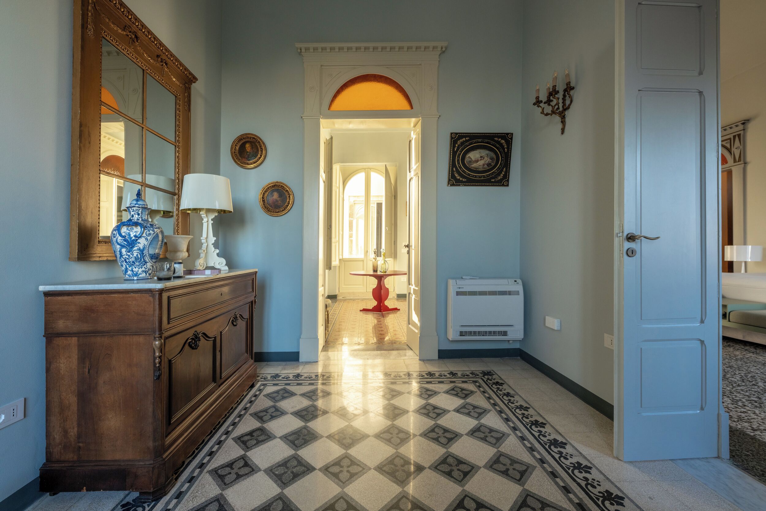 Un couloir avec un sol à motifs, une commode en bois avec un miroir, une table rouge et un appareil de climatisation blanc.