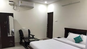 A hotel room with a bed, a desk, a chair, a wardrobe, and an air conditioning unit. - Sri Kamakshi Bhavan (Narayanavanam)