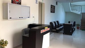 A reception desk with a sign reading "Sri Kamakshi Bhavan," a framed religious icon on the wall, a staircase, and a seating area with chairs. - Sri Kamakshi Bhavan (Narayanavanam)