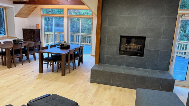 A spacious living area with a fireplace, wooden dining table, and large windows offering a view of the outdoors.