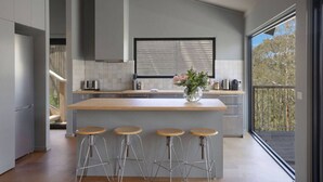 Une cuisine moderne dotée d’un îlot central, de tabourets de bar, d’une fenêtre avec stores et d’une vue sur l’extérieur.