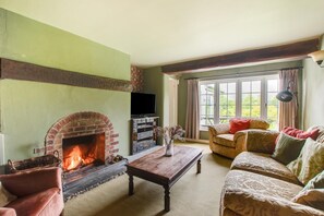 A living room with a brick fireplace, a television, a sofa, and a window with curtains. - 4 Bed in Belper (oc-dcwod) (Paddocks)