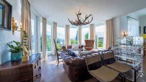 A spacious living room with leather sofas, a glass coffee table, and a chandelier. Large windows offer a view of the outdoors. - Villa Victoria | Sauna | Parking | Garden (Fischerndorf)