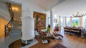A spacious living room with a white decorative column, a staircase with ornate metal railings, a large framed floral painting, and a view of greenery through large windows. - Villa Victoria | Sauna | Parking | Garden (Fischerndorf)
