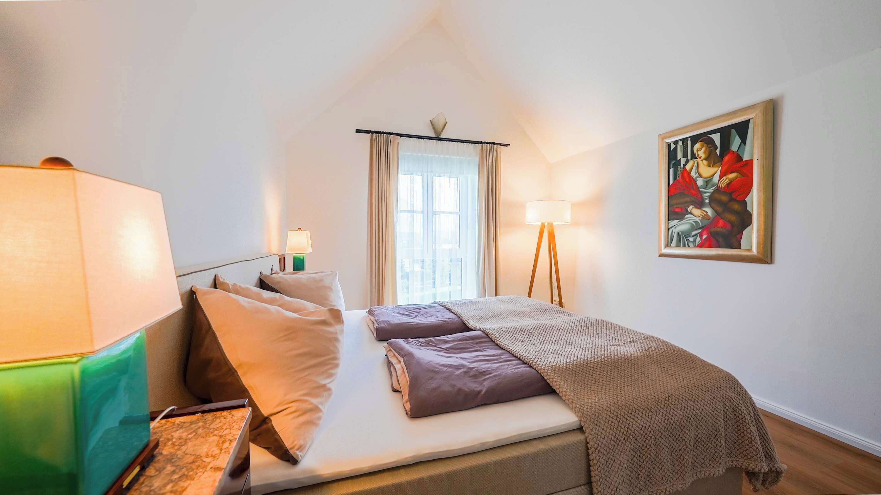 Ein Schlafzimmer mit einem Bett, Nachttischlampen, einem Gemälde und einem Fenster mit Vorhängen.