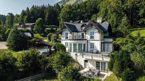 A large, two-story house with a balcony, surrounded by lush greenery and a hilly landscape. - Villa Victoria | Sauna | Parking | Garden (Fischerndorf)
