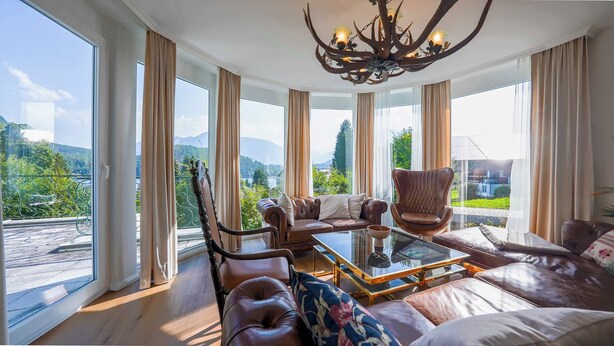 A spacious living room with a large window offering a mountain view, leather furniture, and a chandelier. - Villa Victoria | Sauna | Parking | Garden (Fischerndorf)