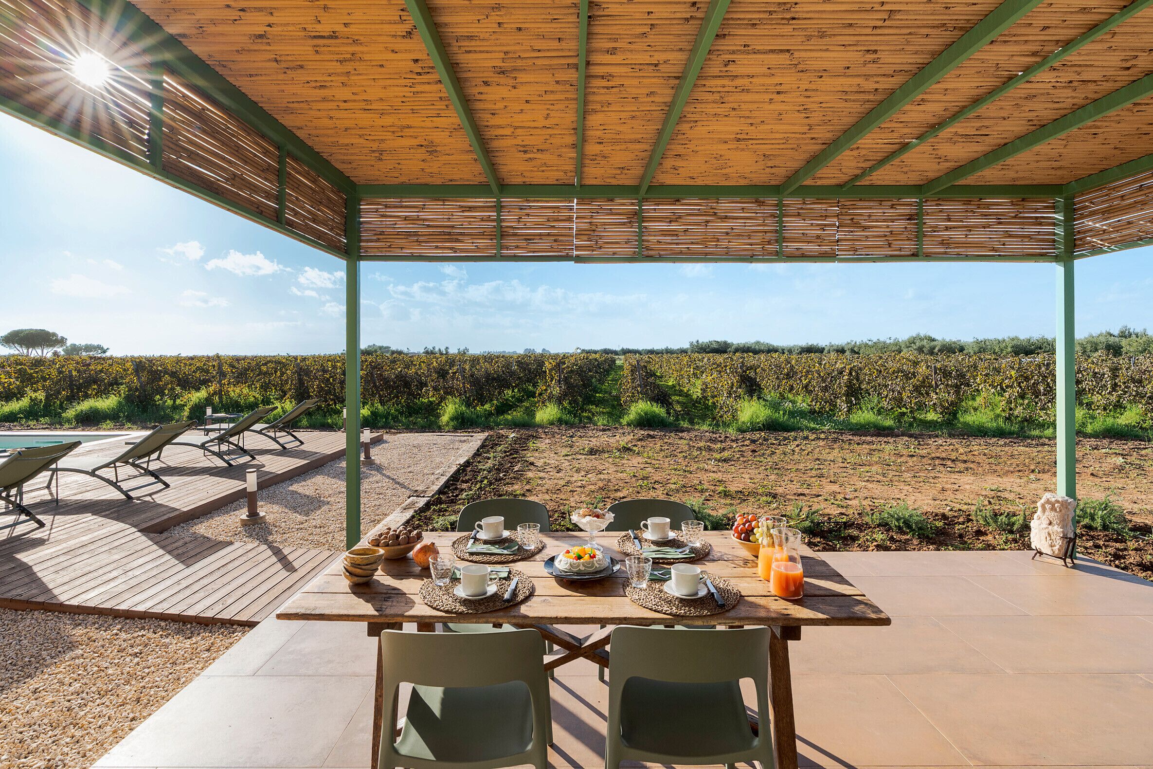 Un espace de restauration en plein air avec une table dressée pour deux personnes, entourée de chaises, sous une pergola en bois offrant une vue sur un vignoble.
