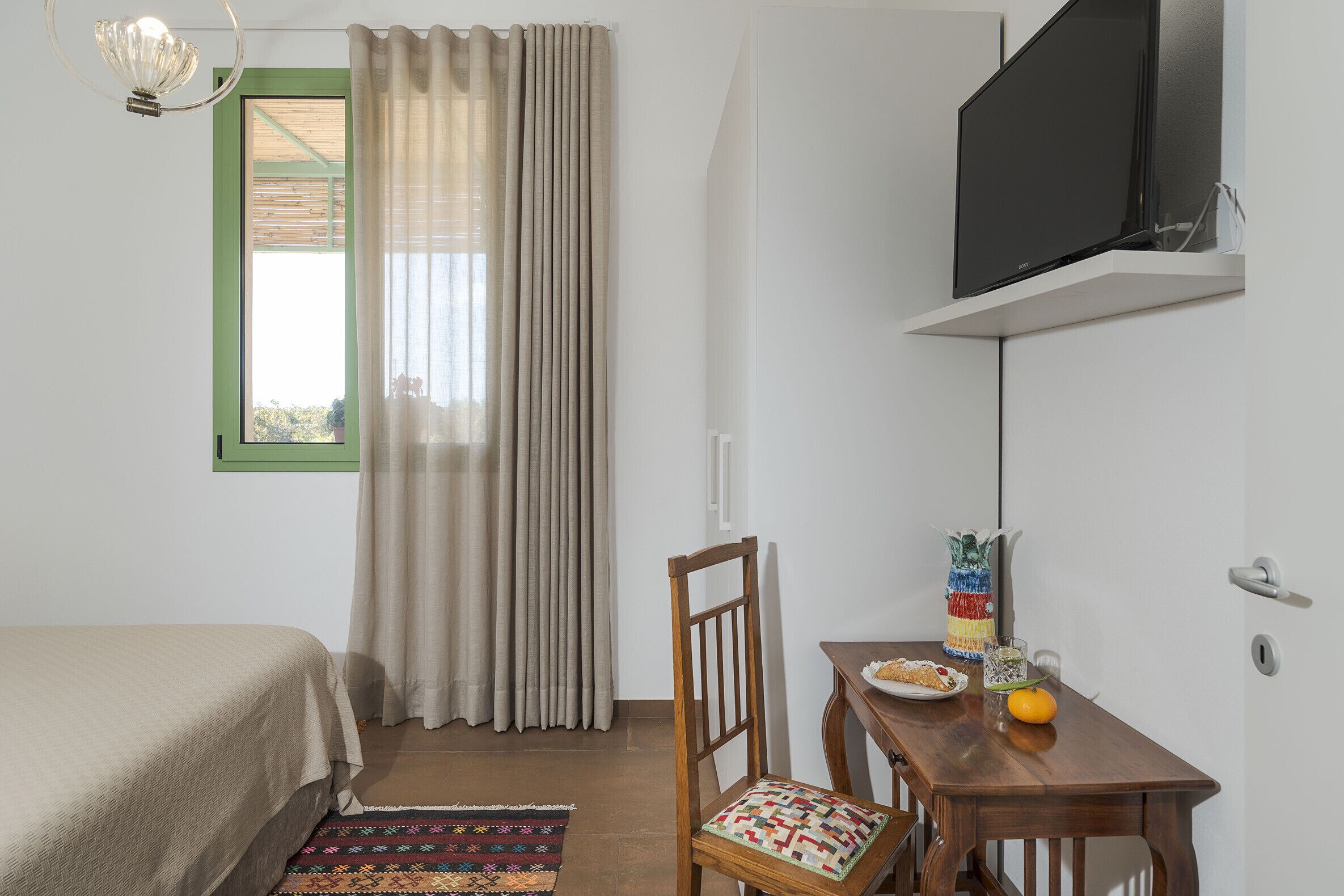A bedroom with a bed, a wooden chair, a small table with a plate of food, a vase, and a television on a stand.
