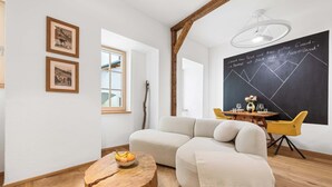 Un salon moderne avec un canapé d’angle, une table basse en bois sur laquelle sont disposés des fruits, un coin repas avec des chaises jaunes et un tableau noir avec du texte.