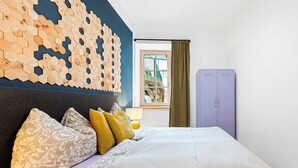 Un lit bien fait, agrémenté d’oreillers à motifs, une décoration murale en bois de forme hexagonale, une fenêtre avec des rideaux et une armoire violette.