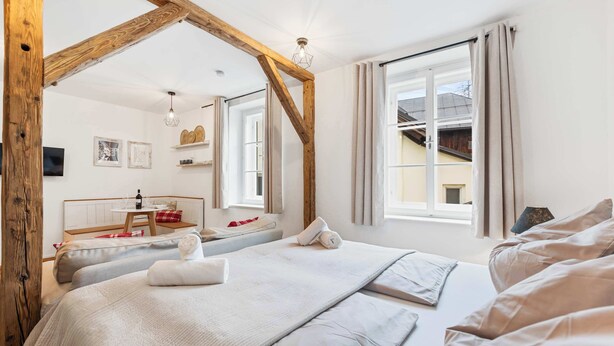 A hotel room with a large bed, a wooden ceiling beam, a window with curtains, and a small table with a red and white checkered tablecloth. - Superior Studio | Kitchen | Parking | Central (Bad Aussee)