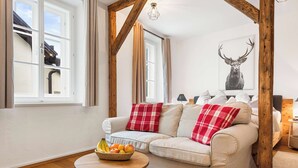 A cozy living room with a beige sofa, red and white checkered throw pillows, a wooden coffee table with a fruit bowl, and a deer artwork on the wall. - Superior Studio | Kitchen | Parking | Central (Bad Aussee)